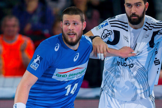 Zwei männliche Handballspieler sind auf dem Spielfeld in Aktion. Der Spieler in Blau, der die Nummer 14 trägt, wird von einem Spieler in einem weißen Trikot eng bewacht. Beide tragen Bärte und scheinen während des Spiels konzentriert zu sein.