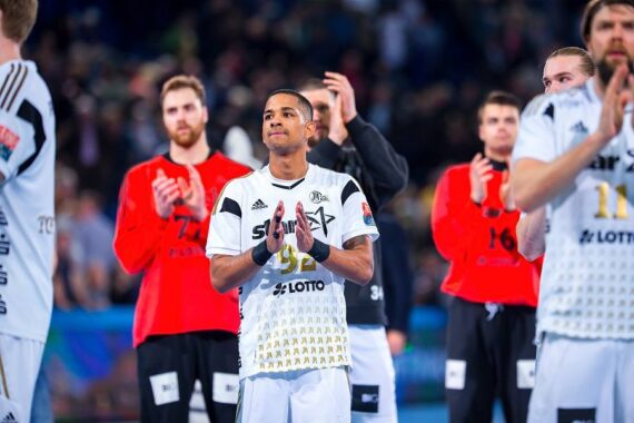 Ein männlicher Handballspieler in einem weißen Trikot steht im Vordergrund und klatscht, während seine Mannschaftskameraden und Spieler in roten Trikots im Hintergrund in einer hell erleuchteten Halle zu sehen sind.