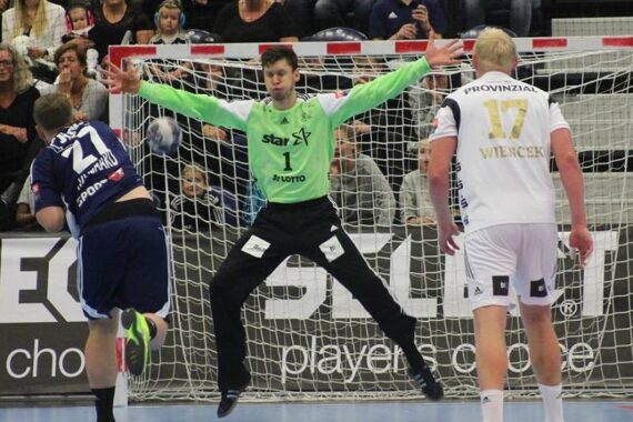 Ein Handballtorwart in einem grünen Trikot springt mit gespreizten Armen und Beinen, um einen Schuss abzublocken, während sich zwei gegnerische Spieler in blau-weißen Trikots auf einem Hallenplatz vor den Augen der Zuschauer gegenüberstehen.