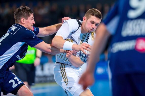 Zwei männliche Handballspieler in Aktion; der eine in einem weißen Trikot hält den Ball, während er von einem Spieler in einem dunkelblauen Trikot während eines intensiven Spiels eng verteidigt und festgehalten wird.