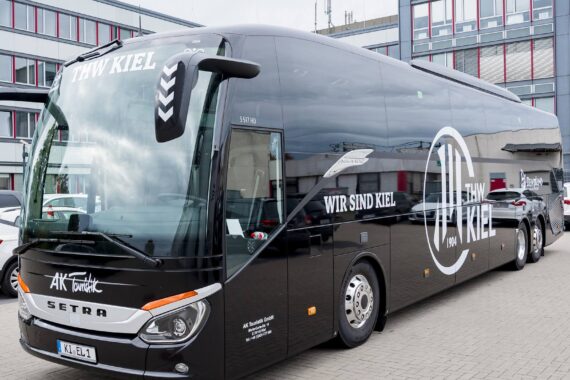 Ein großer schwarzer Setra-Bus mit dem Schriftzug THW Kiel und Wir sind Kiel auf der Seite parkt vor einem modernen Gebäude mit roten und grauen Akzenten.