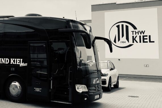 Ein schwarzer Bus mit der Aufschrift WIR SIND KIEL parkt neben einem weißen Auto vor einem Gebäude mit einem großen THW KIEL 1904-Logo an der Wand. Das Bild ist in schwarz-weiß gehalten.