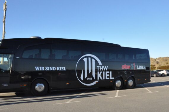 Ein schwarzer Tourbus mit dem Logo des THW Kiel und dem Schriftzug WIR SIND KIEL, der an einem sonnigen Tag auf einem Parkplatz geparkt ist, mit ein paar Autos und felsigem Gelände im Hintergrund.