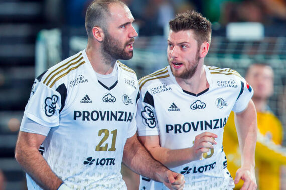 Zwei männliche Handballspieler in weißen Trikots mit goldenen Akzenten und der Aufschrift PROVINZIA stehen dicht beieinander auf einem Spielfeld und wirken konzentriert und engagiert. Im Hintergrund ist ein verschwommener Torwart zu sehen.