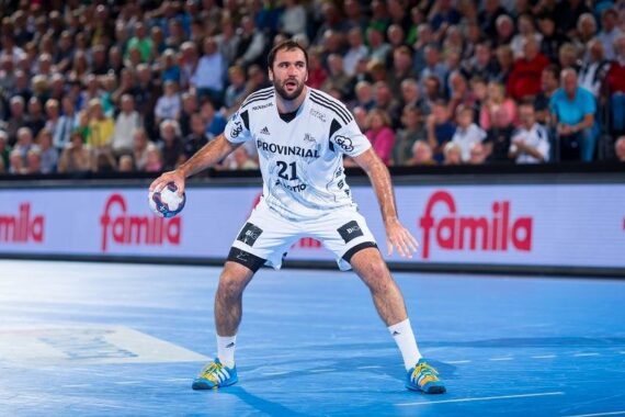 Ein männlicher Handballspieler in einem weißen Trikot steht auf einem blauen Spielfeld bereit und hält mit beiden Händen einen Ball. Ein Publikum schaut von der Tribüne im Hintergrund zu.