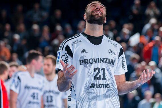 Ein Handballspieler, der ein weißes Trikot mit der Nummer 21 trägt, steht auf dem Spielfeld, blickt auf und hebt die Hände, wirkt emotional. Im Hintergrund sind andere Spieler und ein verschwommenes Publikum zu sehen.