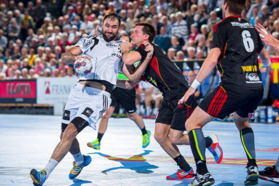 Ein Handballspieler in einem weißen Trikot versucht, mit dem Ball vorzurücken, während ein Verteidiger in einem schwarzen Trikot ihn blockiert; im Hintergrund sind zwei weitere Spieler und eine große Menschenmenge zu sehen.