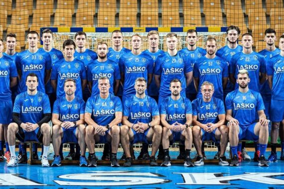 Eine Gruppe von Männern in blauen Sportuniformen posiert für ein Mannschaftsfoto vor einem Hallennetz; die meisten stehen, einige sitzen, alle tragen Hemden mit der Aufschrift Laško.