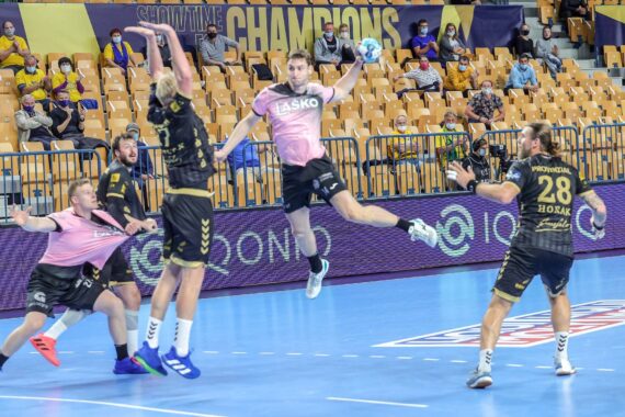 Ein Handballspieler in einem rosafarbenen Trikot springt zum Wurf, während drei Gegner in schwarzen Trikots den Ball verteidigen, während die Zuschauer in gelben Sitzen im Hintergrund zusehen.