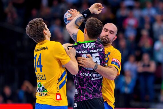 Zwei männliche Handballspieler in gelben Trikots blockieren einen gegnerischen Spieler in einem schwarz-lila Trikot, der den Ball während eines intensiven Spiels hält, mit einer verschwommenen Menschenmenge im Hintergrund.