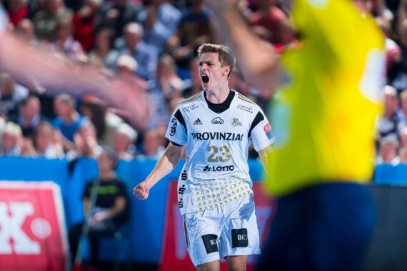 Ein männlicher Handballspieler in einem weißen Trikot mit der Nummer 23 schreit während eines Spiels leidenschaftlich, mit einem unscharfen Schiedsrichter in Gelb im Vordergrund und einer Menschenmenge im Hintergrund.