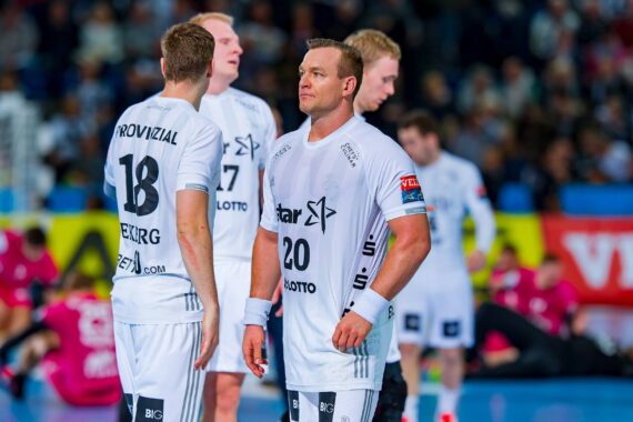 Eine Gruppe von männlichen Handballspielern in weißen Trikots steht auf einem Hallenplatz zusammen. Der vorderste Spieler blickt nach vorne, während sich andere in der Nähe unterhalten. Im Hintergrund sind unscharfe Spieler und Zuschauer zu sehen.