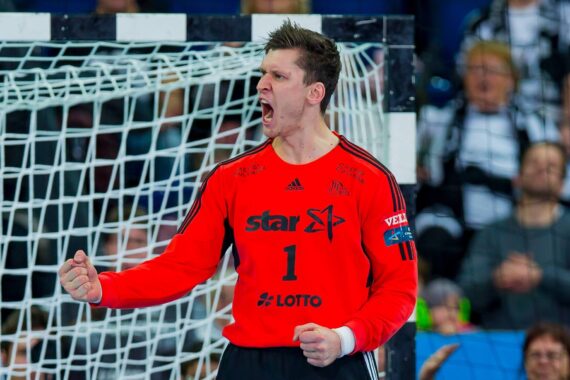 Ein männlicher Handballtorwart in einem roten Trikot mit der Nummer 1 und Sponsorenlogos ballt die Faust und jubelt vor einem Tornetz, während im Hintergrund Zuschauer zu sehen sind.