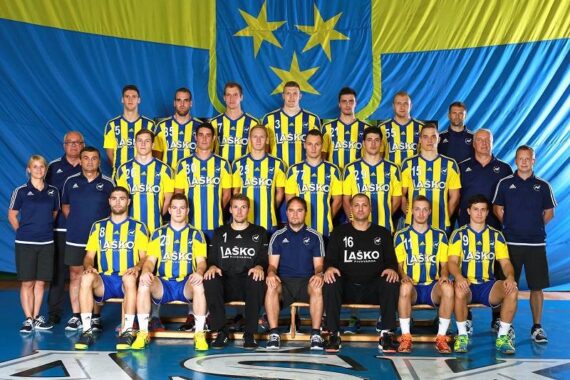 Eine Handballmannschaft der Männer posiert für ein Gruppenfoto in gelb und blau gestreiften Trikots, mit Trainern an den Seiten und einer großen blau-gelben Flagge im Hintergrund.