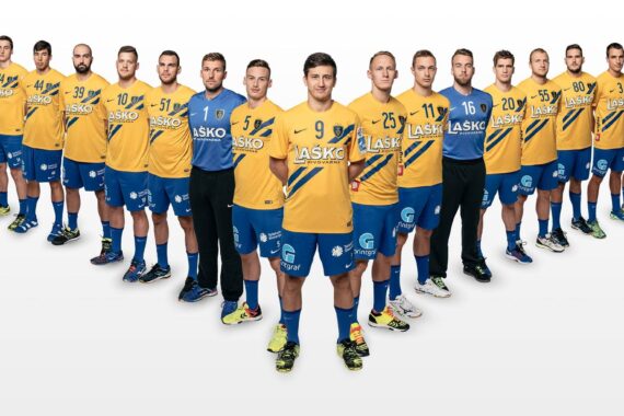 Eine Männer-Handballmannschaft in gelben und blauen Trikots posiert in V-Formation vor einem weißen Hintergrund. Die Spieler stehen in zwei Reihen mit Blickrichtung nach vorne, die Trainer in Schwarz an beiden Enden.