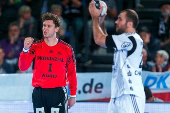Ein Handballtorwart in einem roten Trikot jubelt mit geballter Faust, während ein gegnerischer Spieler in einem weißen Trikot sich auf den Wurf des Balls vorbereitet. Im unscharfen Hintergrund sind Zuschauer zu sehen.