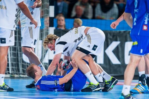Ein Handballspieler in Weiß beugt sich über einen Spieler in Blau, der auf dem Spielfeld liegt und den Ball hält, während andere Spieler während eines intensiven Spiels in der Nähe stehen.