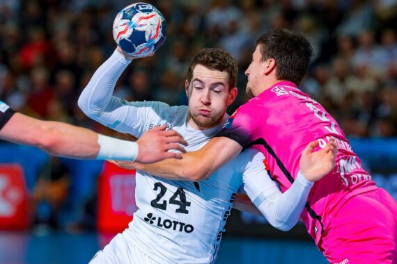 Ein Handballspieler im weißen Trikot (Nummer 24) springt, um den Ball zu werfen, während er während eines Spiels von einem Gegner im rosa Trikot eng verteidigt wird. Die Aktion ist intensiv, beide Spieler sind in Bewegung.