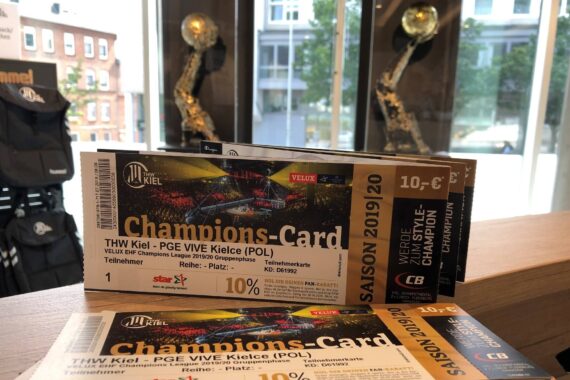 Zwei Champions-Card-Tickets für ein Handballspiel zwischen dem THW Kiel und PGE VIVE Kielce liegen auf einem Tresen aus, im Hintergrund stehen Trophäen und große Fenster.