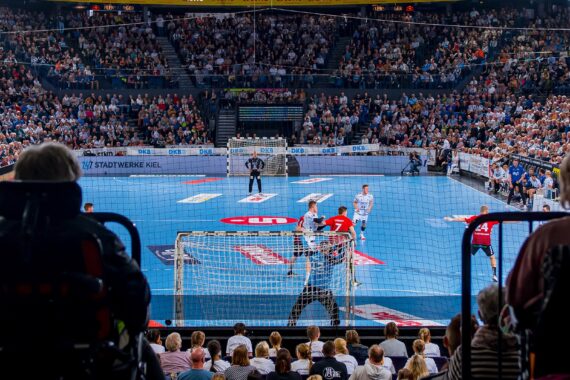 Blick von hinten auf zwei Rollstuhlfahrer, die ein Hallenhandballspiel verfolgen; die Spieler sind auf einem blauen Spielfeld in Aktion, und die Halle ist mit zahlreichen Zuschauern gefüllt.