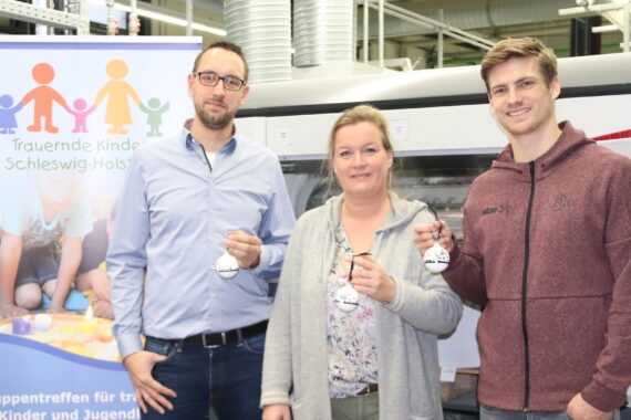 Drei Personen stehen in einem Haus, halten Ornamente und lächeln in die Kamera. Hinter ihnen ist ein buntes Banner mit kindlichen Figuren und deutschem Text für eine Kinderhilfsgruppe zu sehen. Im Hintergrund ist eine Industrieanlage zu sehen.