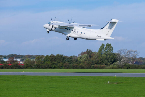 Ein weißes Flugzeug mit zwei Propellern hebt von einer Landebahn ab, im Hintergrund grünes Gras und Bäume unter einem blauen Himmel.