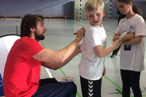 Ein Mann in einem roten Hemd sitzt in einer Turnhalle und unterschreibt auf der Rückseite eines weißen Jungen-T-Shirts, während ein Mädchen mit verschränkten Armen in der Nähe steht und das Geschehen beobachtet.