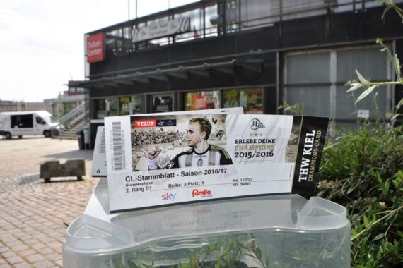 Eine Eintrittskarte für ein Handballspiel des THW Kiel in der Saison 2016/17 ist im Freien ausgestellt. Im Hintergrund ist ein Gebäude mit dem Schild des Ticket Centers und Kopfsteinpflaster zu sehen.