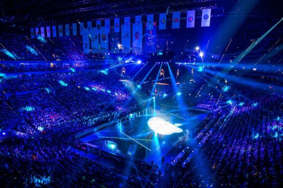 Eine mit Zuschauern gefüllte Arena, die von blauen Lichtern und Scheinwerfern beleuchtet wird, umgibt eine große zentrale Bühne mit von der Decke hängenden Bildschirmen und Bannern, die eine energiegeladene High-Tech-Atmosphäre schaffen.