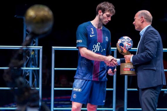Ein männlicher Handballspieler im blauen PSG-Trikot nimmt während einer Preisverleihung einen Pokal von einem Mann im Anzug entgegen, wobei eine weitere Trophäe im Vordergrund unscharf zu erkennen ist.