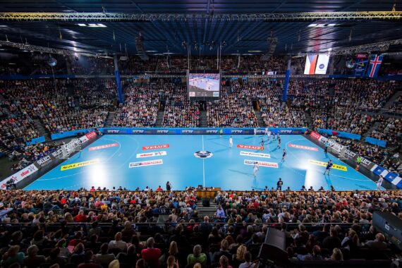 Eine große Halle, gefüllt mit Zuschauern, die ein Handballspiel auf einem hellblauen Spielfeld verfolgen. Nationale Flaggen und Werbung sind zu sehen, und eine große Anzeigetafel hängt über der Mitte des Spielfelds.