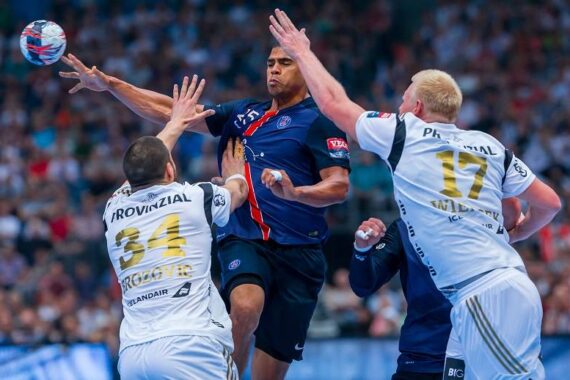 Drei männliche Handballspieler springen und kämpfen um den Ball während eines intensiven Spiels, mit einem Spieler in einem blauen Trikot in der Mitte und zwei Gegnern in weißen Trikots auf beiden Seiten, alle umgeben von einer unscharfen Menge.