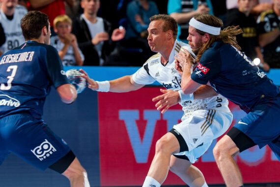 Drei männliche Handballspieler in Aktion während eines Spiels. Ein Spieler in Weiß bewegt sich mit dem Ball vorwärts, während zwei Spieler in Blau versuchen, ihn zu blockieren. Im Hintergrund ist eine Menschenmenge zu sehen, die das intensive Spiel verfolgt.