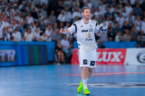 Ein männlicher Handballspieler in einem weißen Trikot hält den Ball in einer Hand und lächelt während eines Spiels auf einem Hallenplatz. Die Zuschauer im Hintergrund sind weiß gekleidet. Der Spieler trägt neongrüne Schuhe.