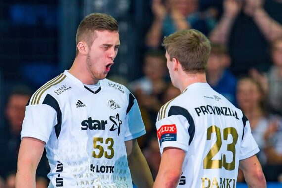 Zwei männliche Handballspieler in weißen Trikots, von denen einer der Kamera zugewandt ist und energisch schreit (#53) und der andere mit dem Rücken zur Kamera steht (#23), scheinen während eines Spiels zu feiern.
