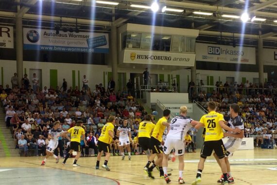 Ein Handballspiel der Männer mit Spielern in gelben und weißen Trikots in Aktion auf einem Hallenplatz, mit einer großen Zuschauermenge im Hintergrund und Fahnen darüber.