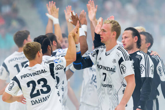 Eine Gruppe männlicher Handballspieler in weißen Trikots klatscht sich auf dem Spielfeld gegenseitig ab und feiert ein Spiel. Die Stimmung ist ausgelassen, einige Spieler schauen in die Kamera, andere wenden sich ab.