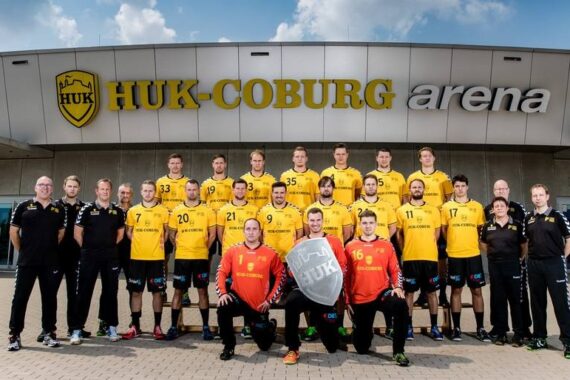 Eine Handballmannschaft in gelben und schwarzen Trikots posiert mit Trainern und Mitarbeitern vor der HUK-Coburg Arena. Zwei Torhüter in roten Trikots sitzen vorne, der Rest steht in Reihen hinter ihnen.