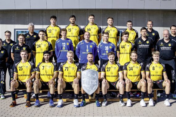Eine Herrenhandballmannschaft und ihr Trainerstab, alle in gelb-schwarzen Trikots, posieren gemeinsam im Freien in einer Reihe. Die Mannschaft hält ein silbernes Schild mit der Aufschrift HSC 2000 vor sich.