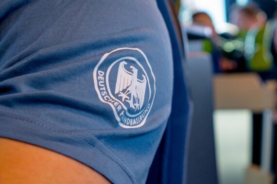 Nahaufnahme des Arms einer Person, die ein blaues Hemd trägt, auf dem das Logo des Deutschen Handballbundes, ein Adler, in weiß aufgedruckt ist.