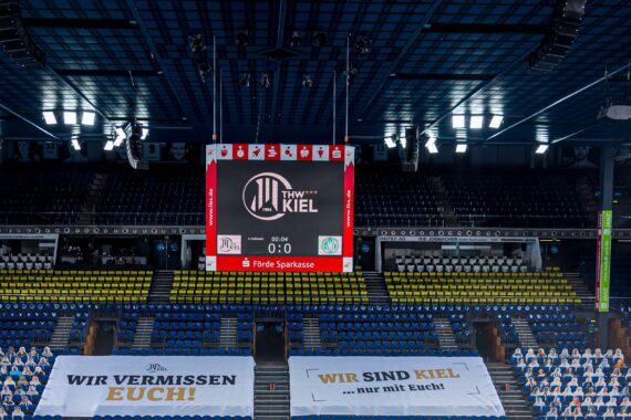 Eine leere Sporthalle mit einer großen Anzeigetafel, die den THW Kiel und ein 0:0 anzeigt. Im Vordergrund sind Banner mit den Aufschriften WIR VERMISSEN EUCH! und WIR SIND KIEL zu sehen, und einige Sitzplätze sind mit Scherenschnitten versehen.