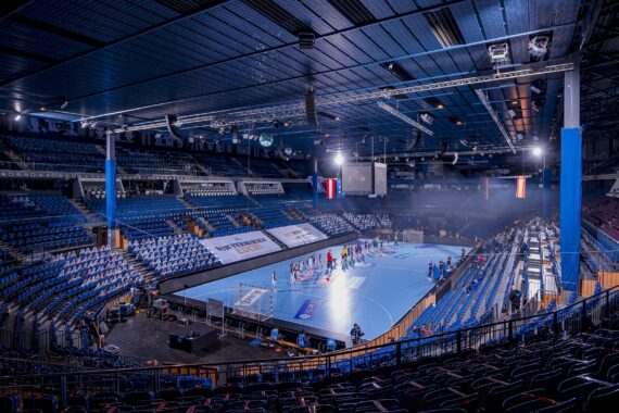 Große Sporthalle mit blauen und lilafarbenen Sitzen, einem blauen Spielfeld in der Mitte und einer Gruppe von Menschen, die auf dem Spielfeld stehen; Fahnen und Scheinwerfer hängen von der Decke, und die Tribünen sind meist leer.