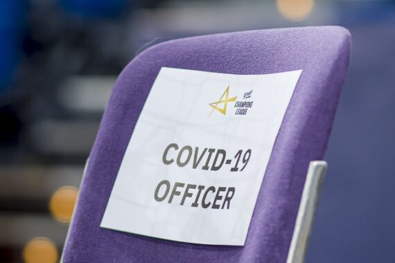Ein violetter Stuhl mit einem Papierschild an der Rückenlehne, auf dem in fetten Buchstaben COVID-19 OFFICER steht. Der Hintergrund ist unscharf.