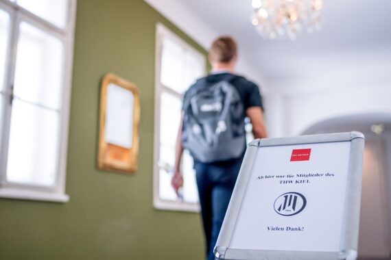 Eine Person mit einem Rucksack geht einen Flur mit grünen Wänden entlang. Im Vordergrund ist ein Schild in deutscher Sprache zu sehen, das den Zugang für Mitglieder des THW Kiel beschränkt. Im Hintergrund sind ein Kronleuchter und ein Spiegel zu sehen.