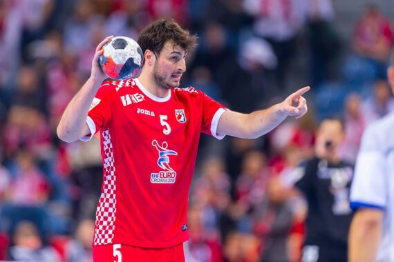 Ein männlicher Handballspieler in einem roten Trikot bereitet sich darauf vor, den Ball zu werfen, während er während eines Spiels nach vorne zeigt. Zuschauer und andere Spieler sind im unscharfen Hintergrund zu sehen.