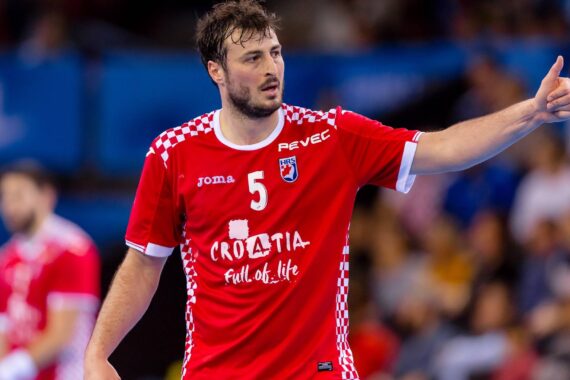 Ein männlicher Handballspieler im roten Kroatien-Trikot mit der Nummer 5 zeigt während eines Spiels den Daumen nach oben, während im Hintergrund eine unscharfe Menschenmenge zu sehen ist.