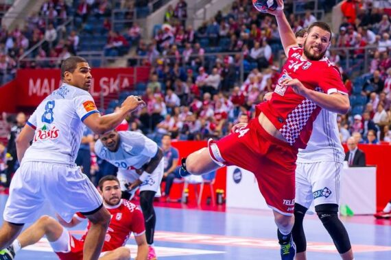 Ein Handballspieler in einem roten Trikot springt, um den Ball zu werfen, während Spieler in weißen Trikots ihn verteidigen. Das Spiel findet in einer Halle statt, im Hintergrund ist eine Menschenmenge zu sehen.