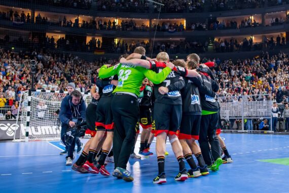 Eine Handballmannschaft steht im Kreis auf dem Spielfeld und jubelt, während im Hintergrund eine jubelnde Menge in einer großen Halle steht.