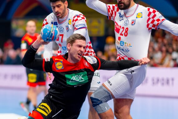 Ein Handballspieler in einem schwarz-roten Trikot versucht zu schießen, während zwei Verteidiger in weiß-rot karierten Trikots versuchen, ihn während eines intensiven Spiels zu blockieren.