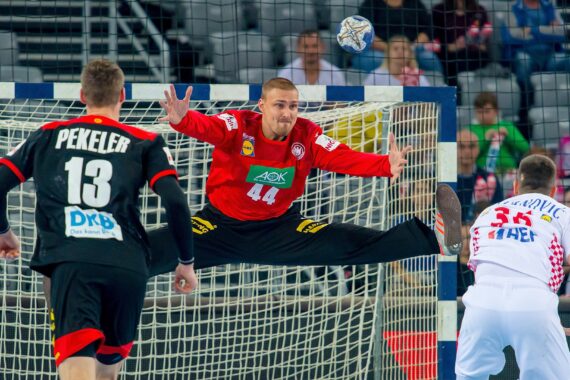 Ein Handballtorwart in einem roten Trikot springt und breitet seine Arme und Beine weit aus, um einen Schuss abzuwehren, während zwei Spieler in schwarz-weißen Trikots auf das Tor zielen. Im Hintergrund sind Zuschauer zu sehen.